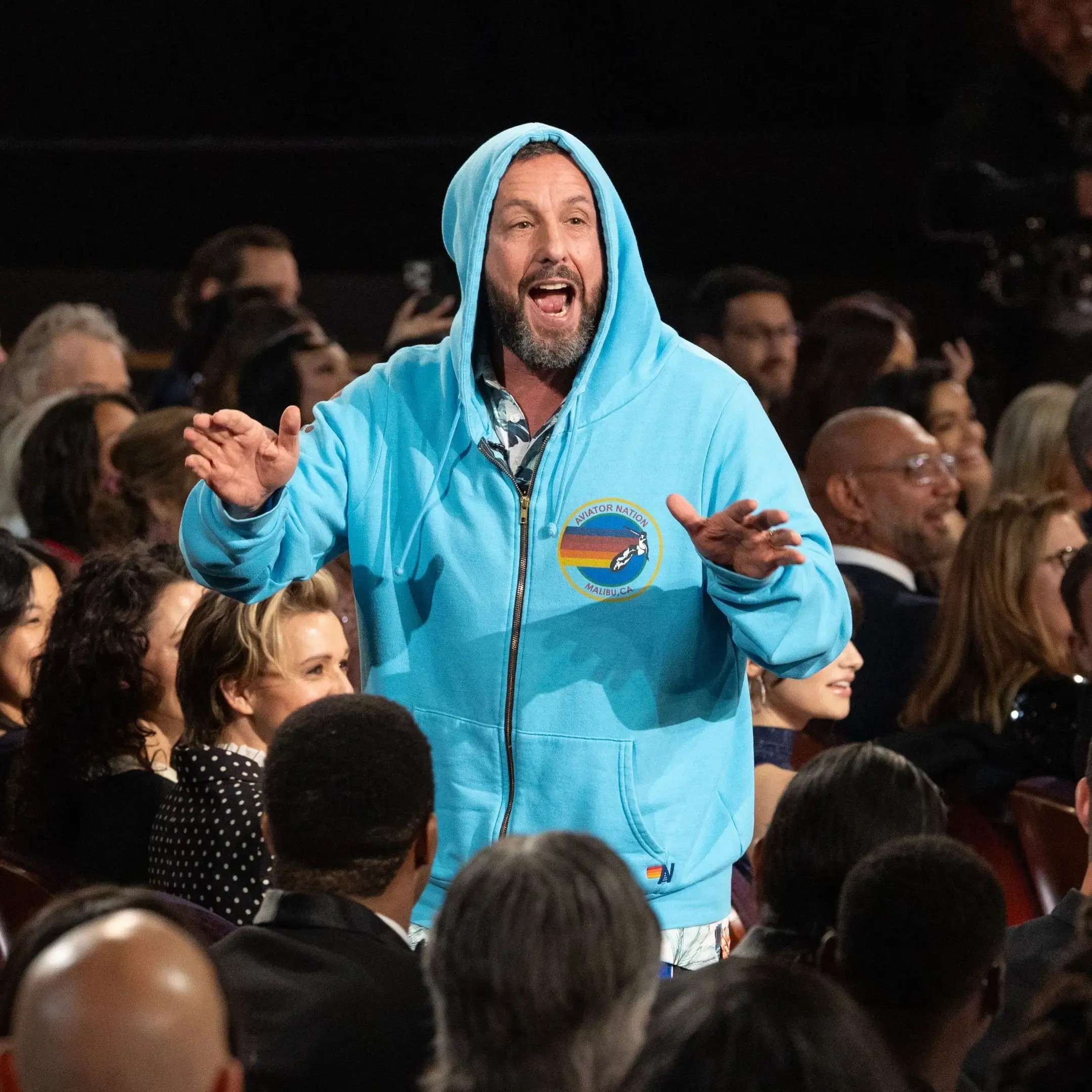 Adam Sandler in a blue hoodie at the Oscars, surrounded by audience members.