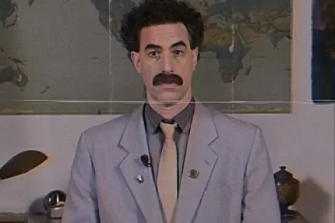 Borat giving a thumbs up in a suit, standing in an office setting.