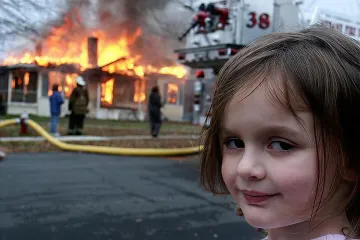 Young girl smirking in foreground with burning house and firefighters in background.