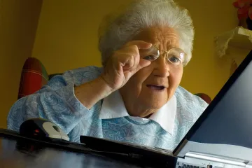 Elderly woman with glasses looking at a laptop screen with curiosity.
