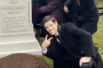 Man in a suit crouching and posing with a peace sign next to a grave.