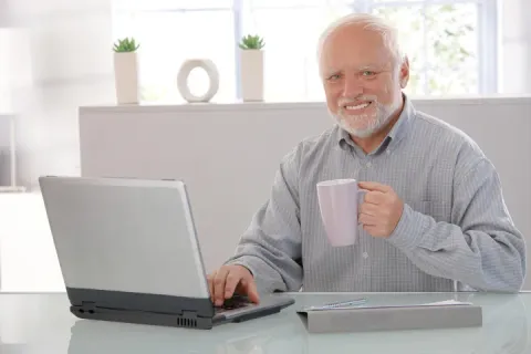 Elderly man with a forced smile holding a mug in front of a laptop.