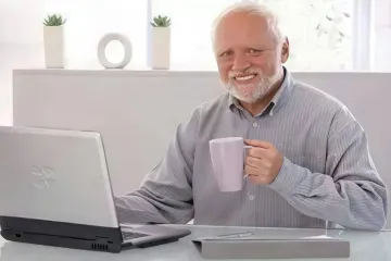 Elderly man with a forced smile holding a mug in front of a laptop.