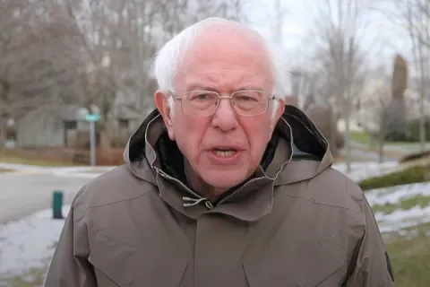 Bernie Sanders in a winter coat speaking outdoors with a serious expression.