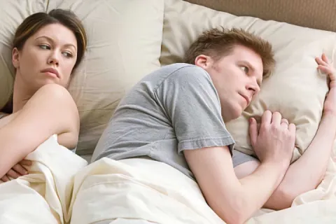 A woman and man lying in bed, woman looking suspiciously at the man.