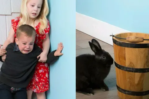 Two children looking scared of a small black rabbit near a wooden bucket.