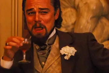 Leonardo DiCaprio in a suit holding a drink, laughing with a scrunched face.