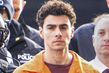 A young man with curly hair, wearing an orange jumpsuit, escorted by law enforcement officers.