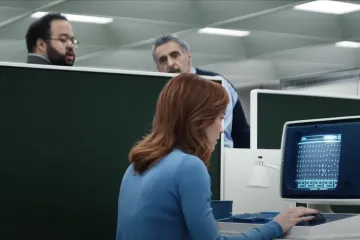 Office scene with three people and a vintage computer screen displaying numbers.