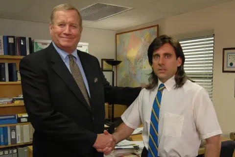 Two men shaking hands in an office setting, one with a mullet hairstyle.