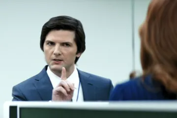 Man in a suit gesturing with finger, talking to a woman in an office setting.
