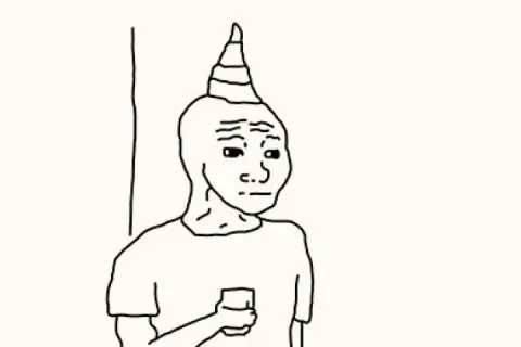 A person with a party hat standing alone at a party, holding a drink, while others dance.