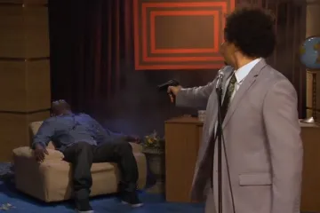 A man in a suit holding a gun with a shocked expression, another man slumped on a chair.