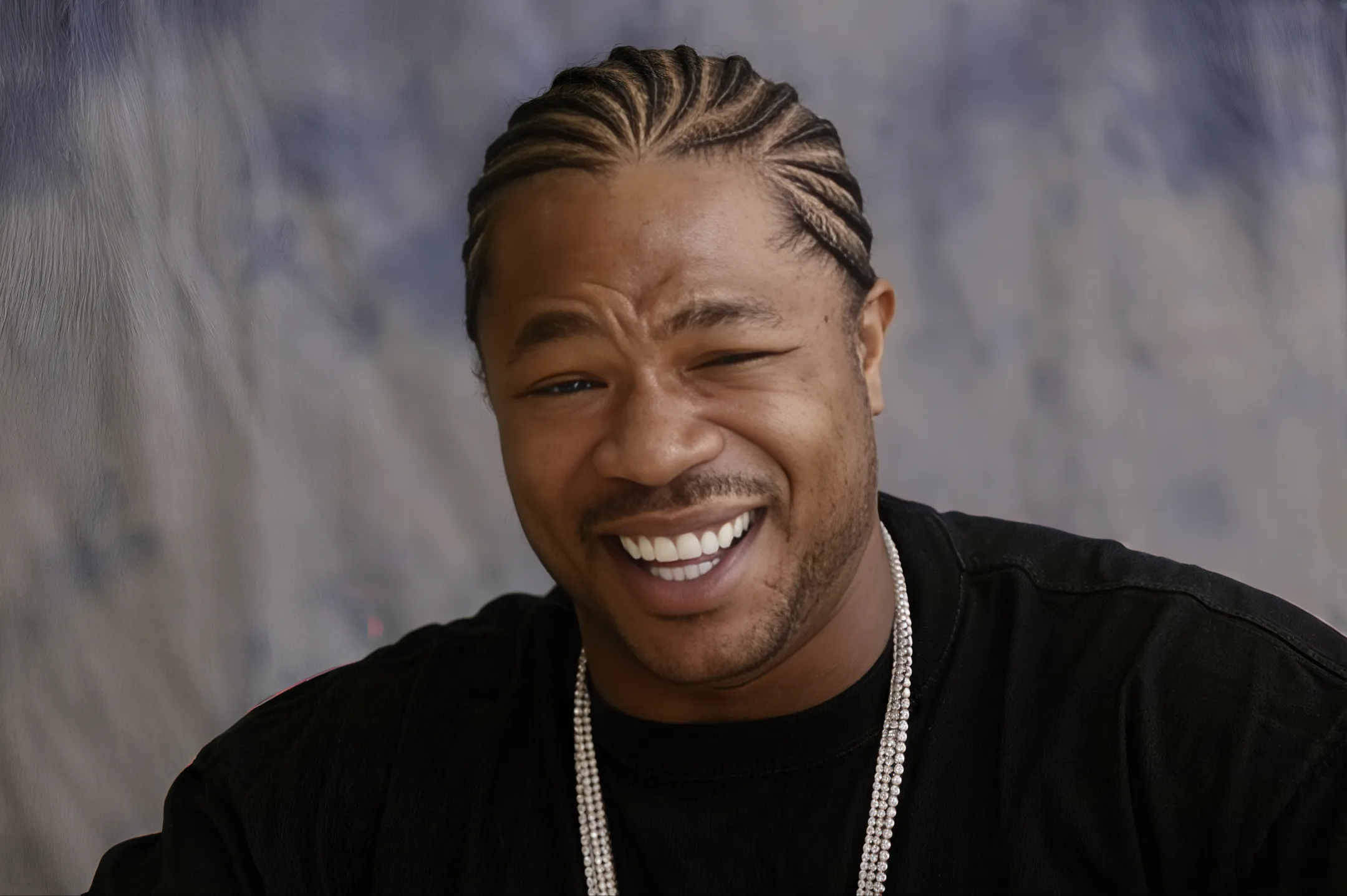 Xzibit smiling with braided hair, wearing a black shirt and chain necklace.