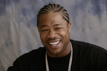 Xzibit smiling with braided hair, wearing a black shirt and chain necklace.